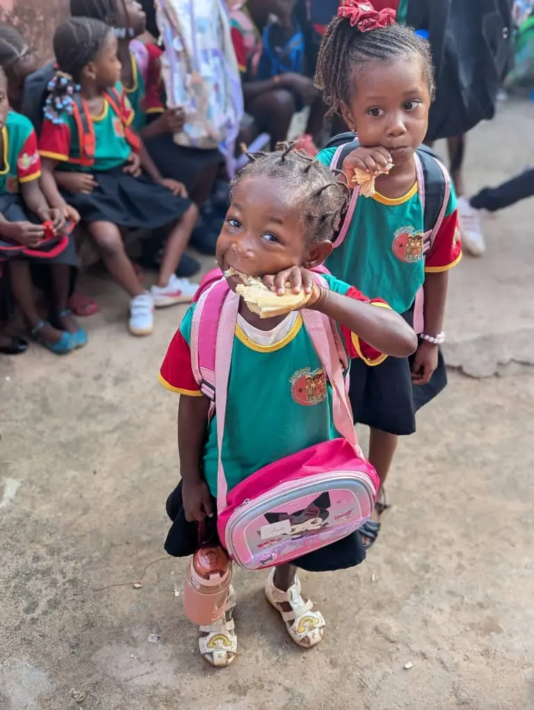 Menina com mochila rosa comendo um pão, ao ar livre