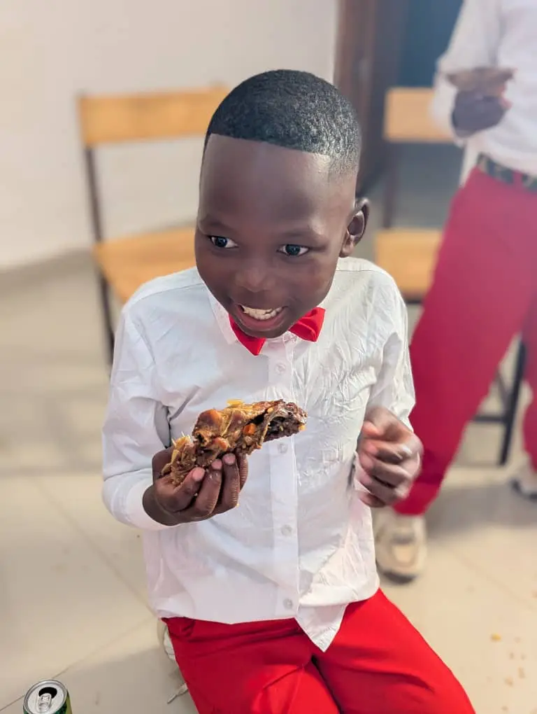 Menino de camisa branca e gravata borboleta vermelha comendo com as mãos, sorrindo