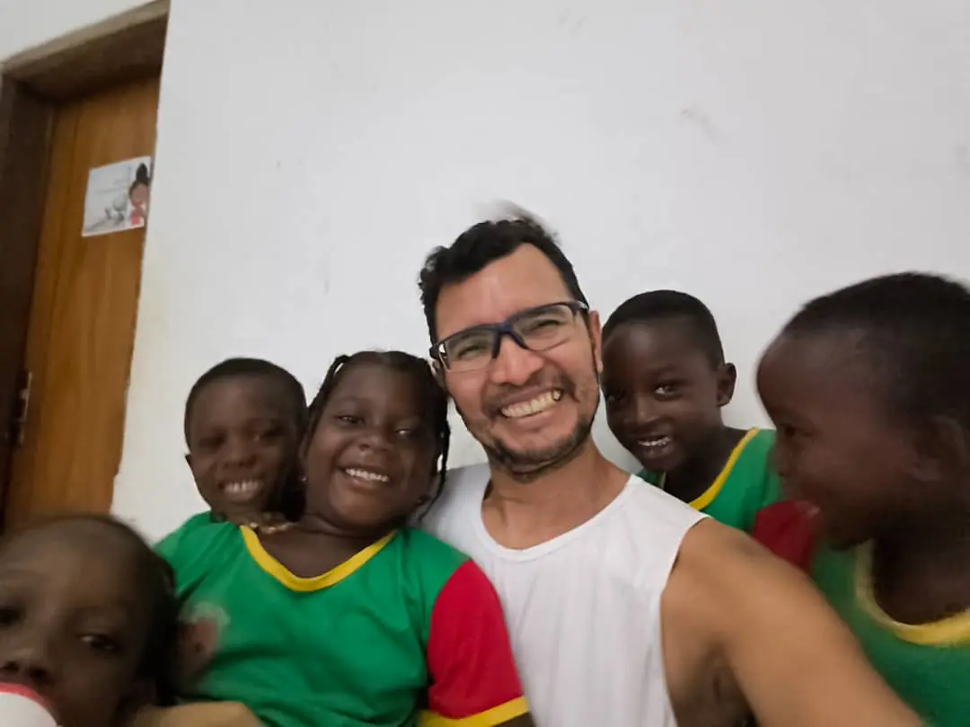 Meninas em fila com os braços cruzados e olhos fechados, em salão amplo